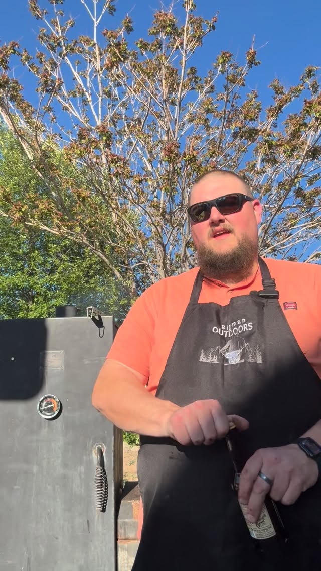 The founder of Bigman Outdoors in a branded apron, tending his smoker outdoors