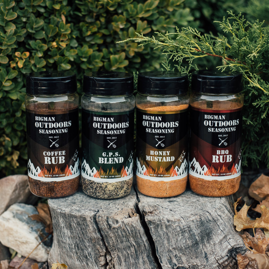 The four Bigman Outdoors rubs — Coffee Rub, G.P.S. Blend, Honey Mustard, and BBQ Rub — lined up on a tree stump outdoors
